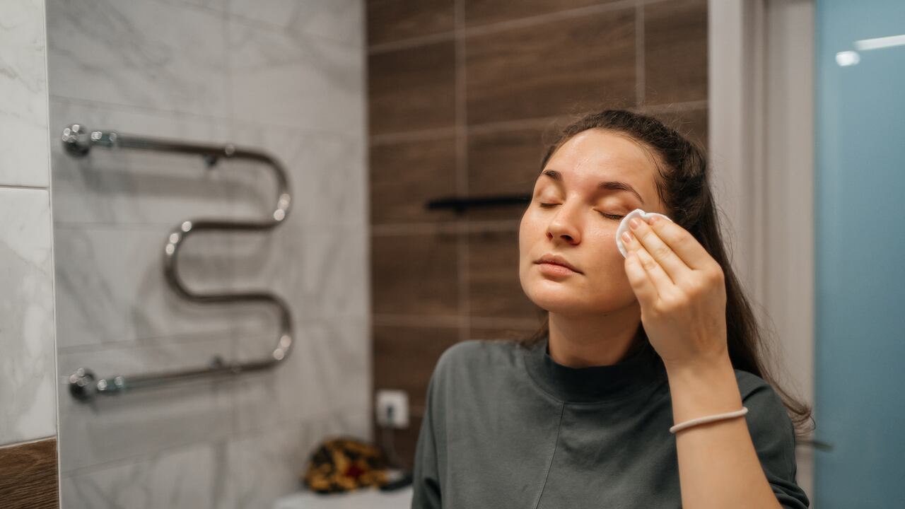 woman wiping face with moisturizing lotion with cotton pad in front of mirror in bathroom at home