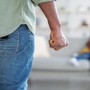 Domestic Violence. Unrecognizable African American Man Threatening Wife And Daughter With His Fist, Scared Mother Embracing Little Girl While Sitting Together On Couch, Selective Focus On Male Hand