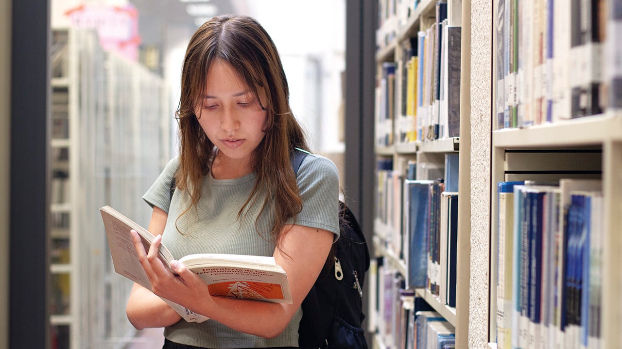 Student international library university bookshelves education. woman looking cheerful returned after reading standing near bookshelves. latin female student holding book literature lesson the break.