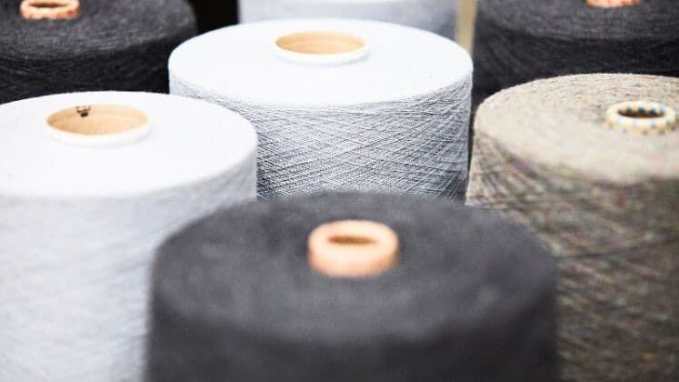 Primer plano de carretes de hilo en tonos neutros, hechos a partir de materiales textiles reciclados, sobre una mesa de trabajo en un taller de moda sostenible.