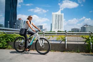 Vista lateral feliz sonriente mujer de negocios de 45 años en bicicleta en bicicleta eléctrica al aire libre en un día soleado de verano en la ciudad europea, edificios de oficinas borrosa en el fondo