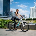 Vista lateral feliz sonriente mujer de negocios de 45 años en bicicleta en bicicleta eléctrica al aire libre en un día soleado de verano en la ciudad europea, edificios de oficinas borrosa en el fondo