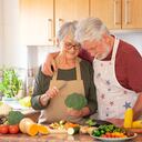 Vegetarian lifestyle. Beautiful smiling senior couple white-haired hug in the kitchen preparing a vegetable soup. On the table a mix of raw seasonal vegetables and a broccoli in the hand