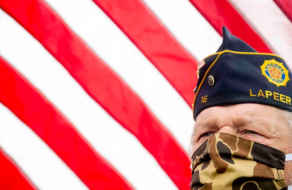 Mike Woodall, de Lum, Mich., que sirvió en el ejército de EE. UU. de 1970 a 1972, sostiene una bandera de EE. UU. mientras usa un tapabocas de camuflaje para protegerse durante un desfile de distanciamiento social y ceremonia del Día de los Caídos, el lunes 25 de mayo de 2020 en Anrook Park in Lapeer, Mich. (Jake May / The Flint Journal vía AP)