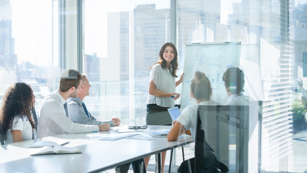Mujer dando una presentación a su equipo. Ella está usando una pizarra con tablas y gráficos. Ella está hablando. Están en una sala de juntas de la oficina en la mesa con computadoras portátiles y papeleo. Ella esta sonriendo. Hombres y mujeres del grupo.