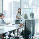 Mujer dando una presentación a su equipo. Ella está usando una pizarra con tablas y gráficos. Ella está hablando. Están en una sala de juntas de la oficina en la mesa con computadoras portátiles y papeleo. Ella esta sonriendo. Hombres y mujeres del grupo.