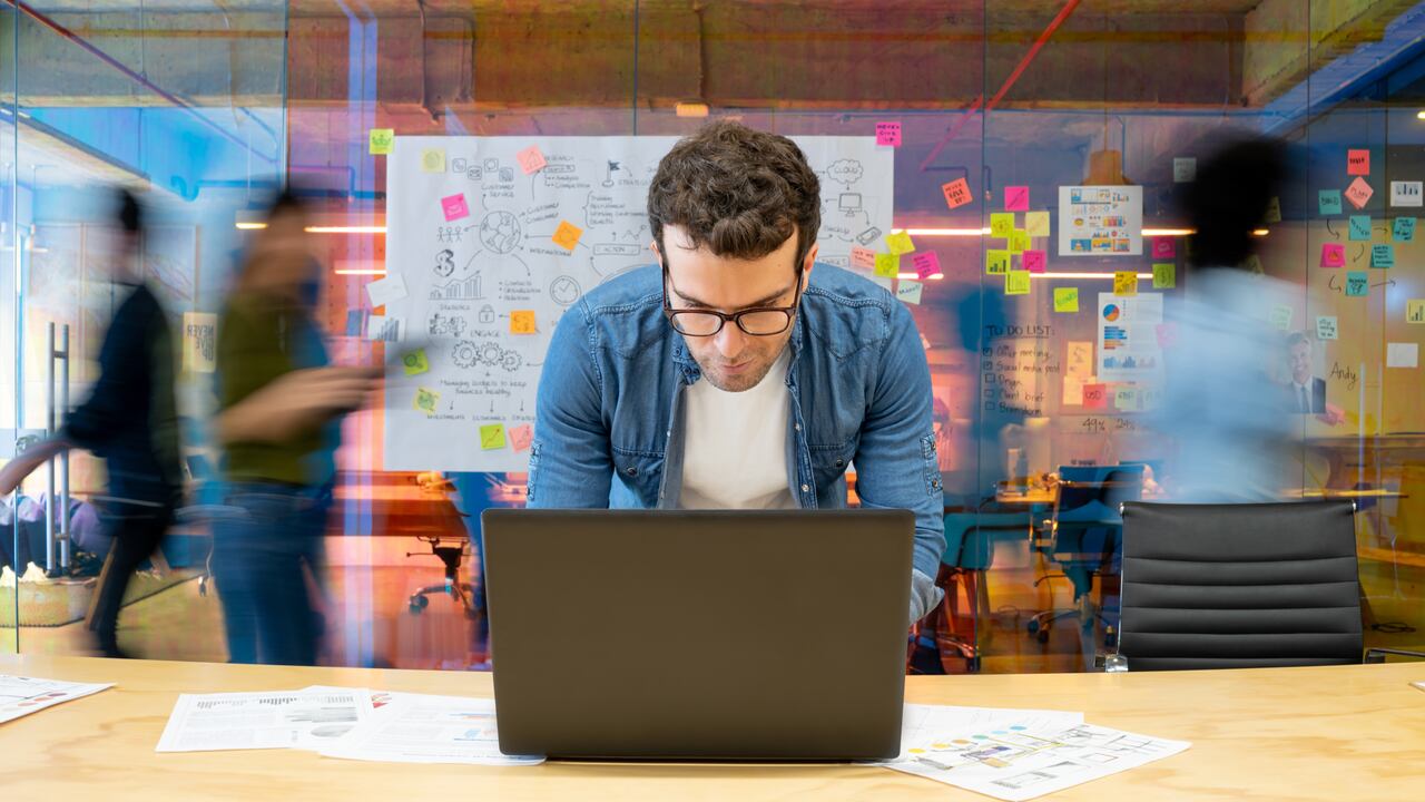 Latin American man working at a creative office using his computer and people moving at the background - place of work concepts