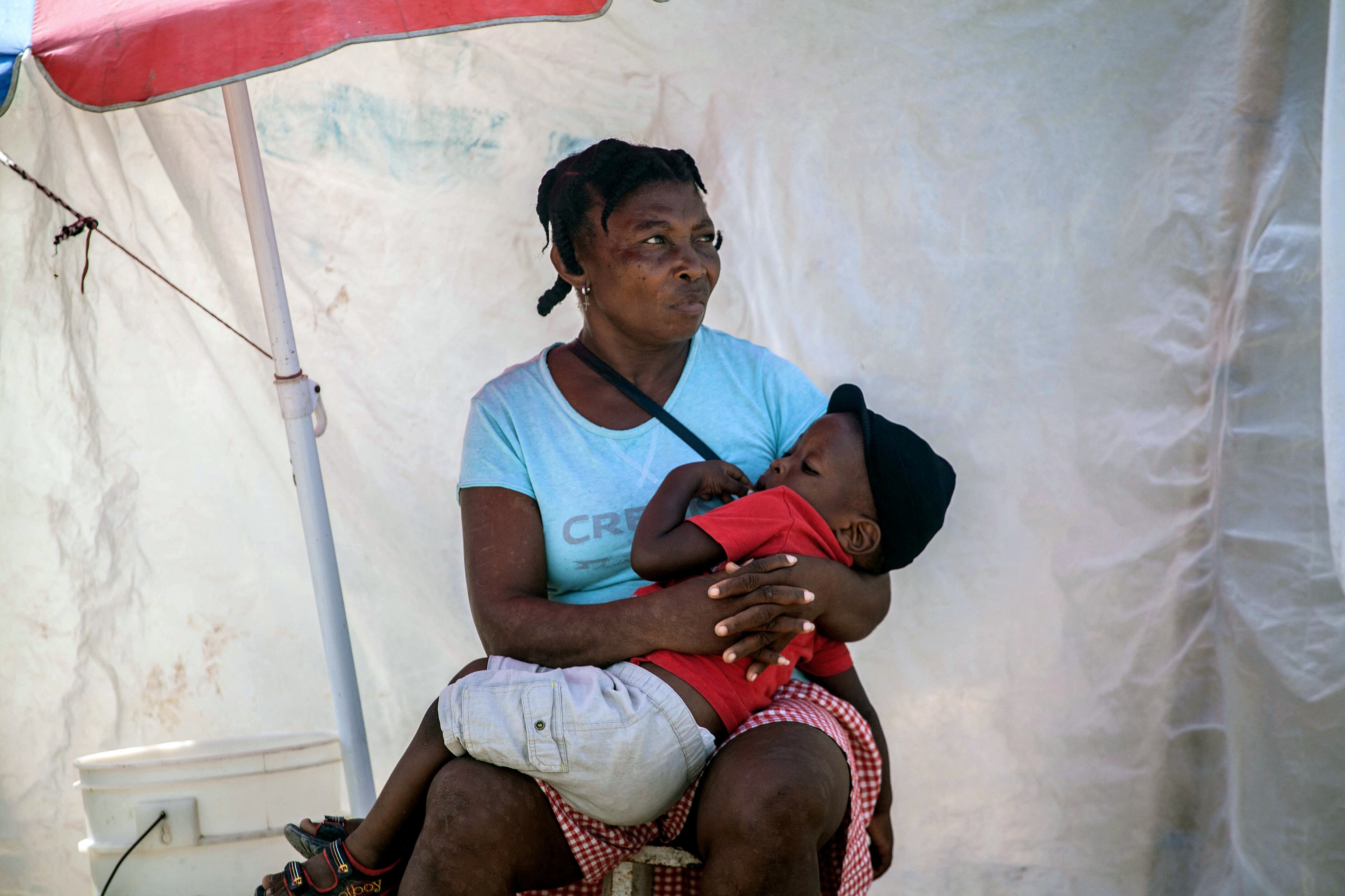A woman holds a boy at a camp for people who lost their home during the August 14 earthquake in Les Cayes, Haiti, on August 23, 2021. - "We're not safe." In a makeshift camp that has served as her refuge since her house collapsed, Vesta Guerrier expresses a fear common to many Haitian women, whom the August 14 earthquake has made extremely vulnerable. Under a flimsy assembly of sheets and plastic sheeting, she lives with her husband and their three children in total destitution on the lawn of a soccer stadium called Gabions in the town of Les Cayes. (Photo by Richard Pierrin / AFP)