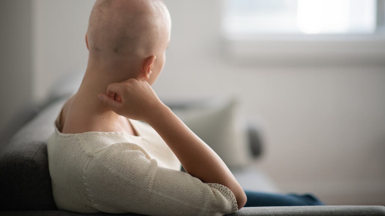 Una mujer está adentro en su sala de estar. Su cabeza está rapada debido a la quimioterapia. Ella está sentada y mirando pensativa