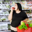 Mujer obesa comiendo una manzana mientras lleva un carrito de compras con verduras orgánicas en el mercado de abarrotes