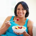 Mujer con sobrepeso sentada en un sofá comiendo un tazón de fruta fresca en casa mirando feliz