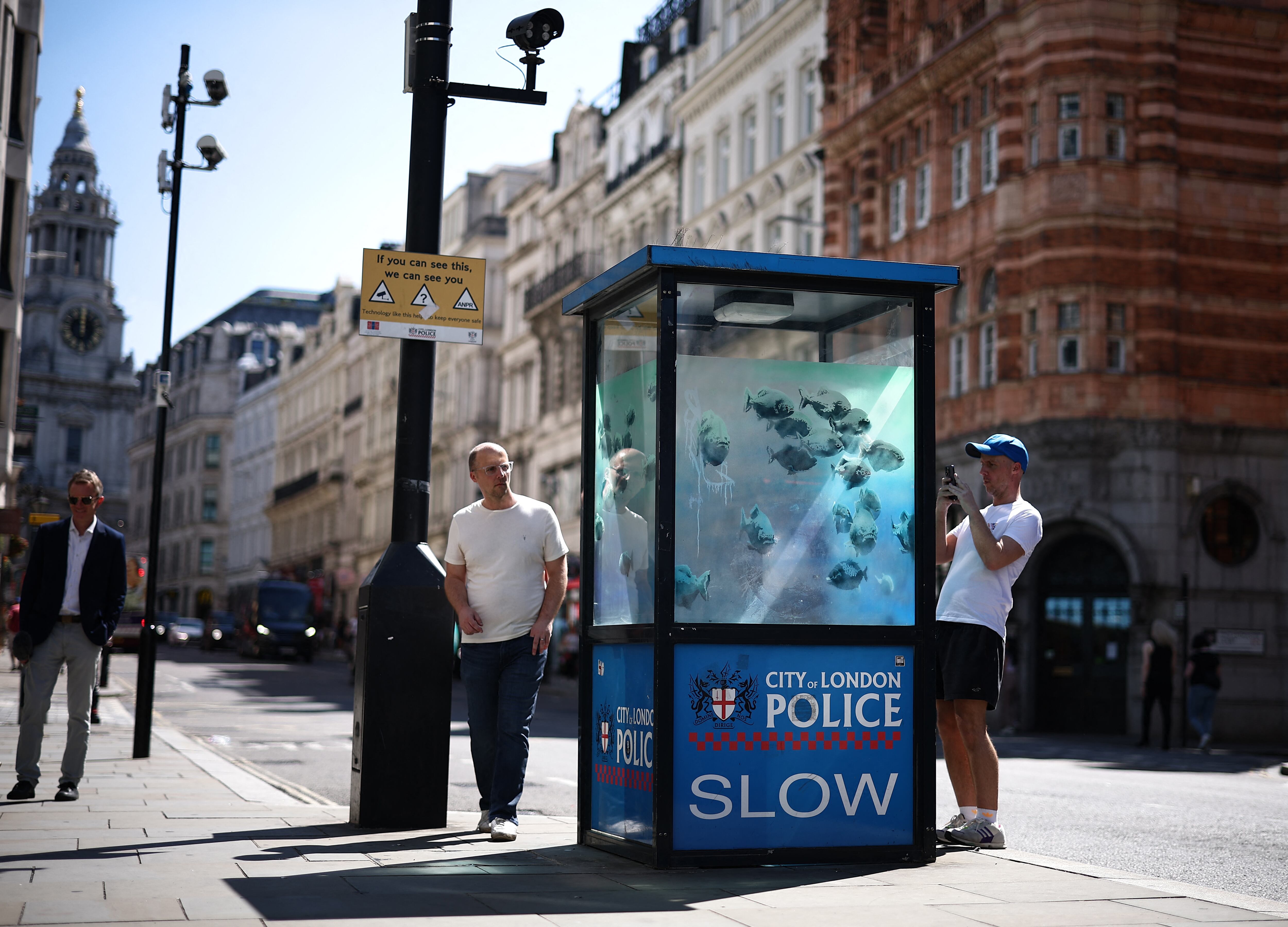 People photograph an artwork by street artist Banksy, the seventh to released this week, depicting fish swimming around a Police Box, in the City of London, on August 11, 2024. The artist confirmed the work to be theirs after posting an image of it on the social media app Instagram. (Photo by HENRY NICHOLLS / AFP) / RESTRICTED TO EDITORIAL USE - MANDATORY MENTION OF THE ARTIST UPON PUBLICATION - TO ILLUSTRATE THE EVENT AS SPECIFIED IN THE CAPTION
