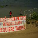Migrants walk past the site where workers are assembling large buoys to be used as a border barrier along the banks of the Rio Grande in Eagle Pass , Texas, Tuesday, July 11, 2023. The floating barrier is being deployed in an effort to block migrants from entering Texas from Mexico. (AP Photo/Eric Gay)