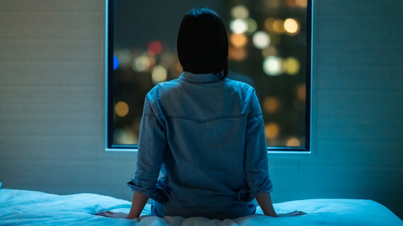 Vista trasera de una mujer sentada sola en la cama en la habitación y mirando por la ventana por la noche