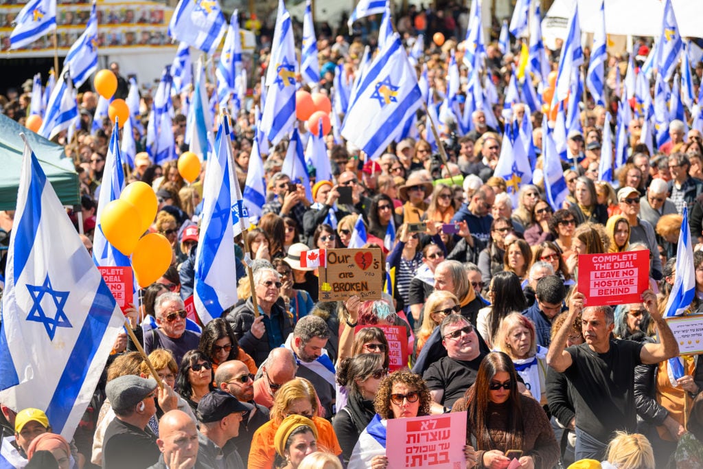 TEL AVIV, ISRAEL - FEBRUARY 26: A crowd watches a live feed from a funeral for members of the Bibas family on a screen in what's known as Hostages Square on February 26, 2025 in Tel Aviv, Israel. A funeral is being held for former hostages Shiri Bibas and her two young sons, Ariel and Kfir, whose bodies were returned to Israel from Gaza last week. At the time of their abduction from Kibbutz Nir Oz on October 7, 2023, Shiri was 32, Ariel was four, and Kfir was nine months old. Their father, Yarden Bibas, 34, was also taken by Hamas militants, but was released alive as part of the ceasefire agreement on 1 February. (Photo by Alexi J. Rosenfeld/Getty Images)