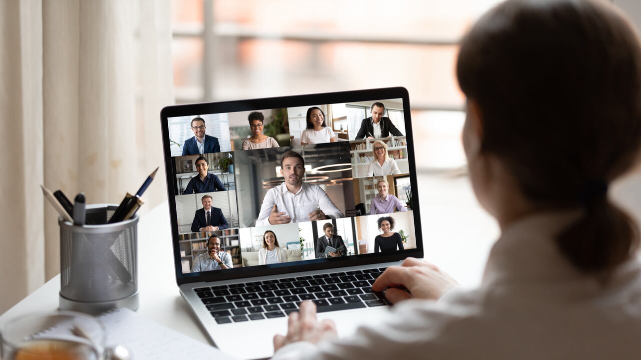 View over businesslady shoulder seated at workplace desk look at computer screen where collage of many diverse people involved at video conference negotiations activity, modern app tech usage concept