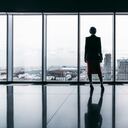 Retrovisor de una empresaria en la parte superior de un rascacielos de oficinas de negocios moderno que se coloca cerca de grandes ventanas panorámicas y que mira pensativamente el paisaje urbano y el sitio de construcción; piso con reflejos