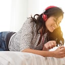 Mujer alegre joven acostada en la cama barajando canciones en el teléfono celular. Chica relajada escuchando música con auriculares en casa. Mujer feliz en casual disfrutando de la música en el dormitorio mientras envía un mensaje de texto por teléfono.