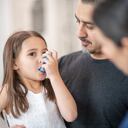 Una doctora muestra a una niña en edad preescolar cómo usar un inhalador para el asma. Ella le está explicando el proceso. La joven está sentada en el regazo de su padre, quien también escucha con atención.