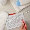 Woman marking critical menstrual days on calendar with pills and intimate care around on white wooden table. Top view.