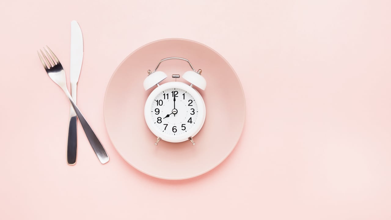 Intermittent fasting concept. White alarm clock on empty pink dish with knife and fork on pink background. Flat lay, copy space for text.