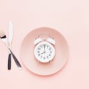 Intermittent fasting concept. White alarm clock on empty pink dish with knife and fork on pink background. Flat lay, copy space for text.