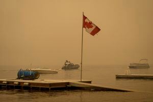 El humo espeso del incendio forestal del lago Lower East Adams llena el aire alrededor de una bandera canadiense ondeando en el viento mientras los oficiales de la Real Policía Montada de Canadá en un barco patrullan el lago Shuswap, en Scotch Creek, Columbia Británica, el domingo 20 de agosto de 2023