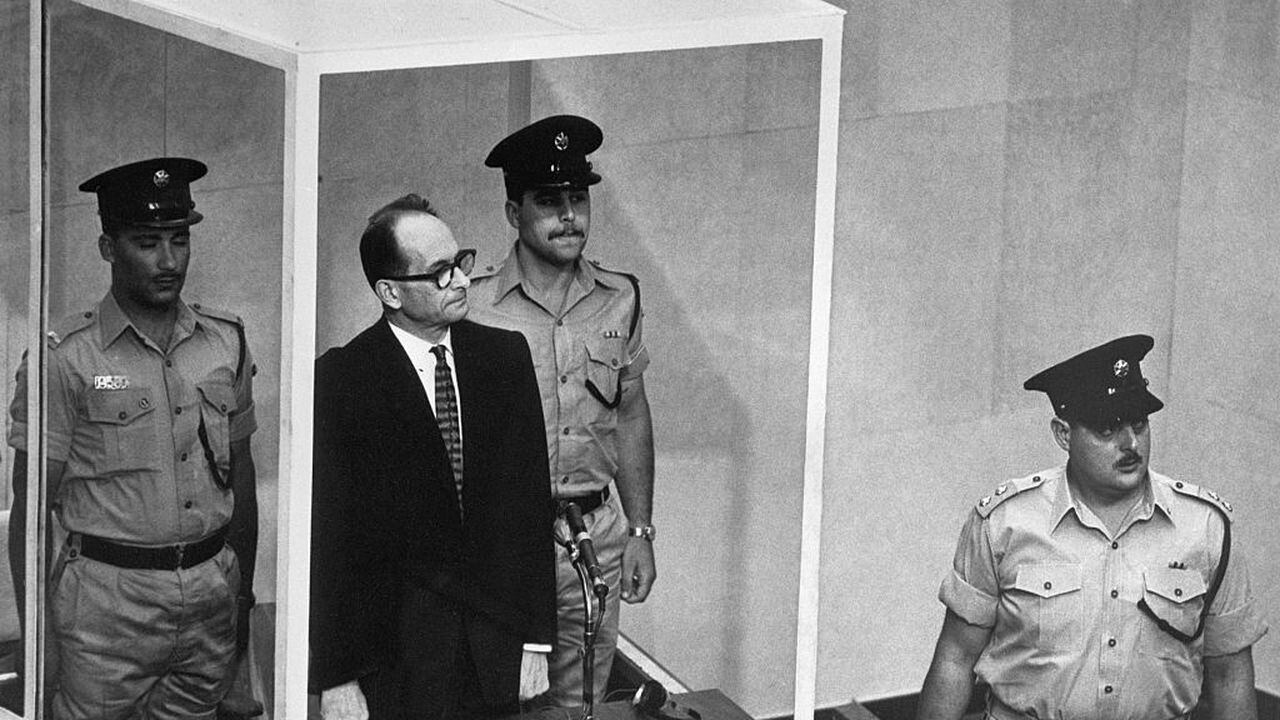 29/5/1962-Jerusalén, Israel- Adolf Eichmann, acusado de asesino en masa nazi, se para en su jaula de vidrio a prueba de balas para escuchar a la Corte Suprema de Israel rechazar por unanimidad una apelación contra su sentencia de muerte. Con él hay dos guardias armados. El veredicto significó que solo el presidente israelí Itzhak Ben-Zvi podía salvar a Eichmann, de 56 años, acusado del asesinato de seis millones de judíos, de la horca. En primer plano está el abogado defensor Robert Servatius.