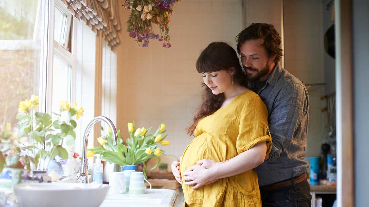 A pregnant couple embrace with there hands gently on stomach in the kitchen of their home with fresh spring flowers in vase.