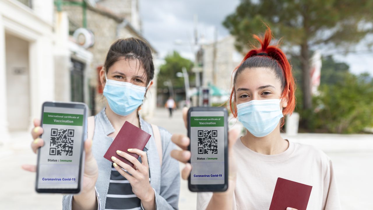 Dos mujeres jóvenes, turista caminando por una ciudad costera con máscaras en el rostro y pasaportes en la mano. Están mostrando una pantalla de teléfono inteligente con certificado electrónico de vacunación COVID19. Unas vacaciones tan esperadas