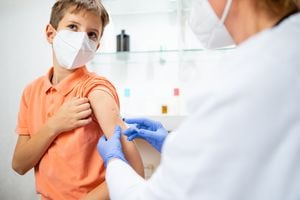 Niño con mascarilla protectora sosteniendo la manga de su camisa y mirando hacia otro lado para no mirar al médico inyectando la vacuna en su brazo
