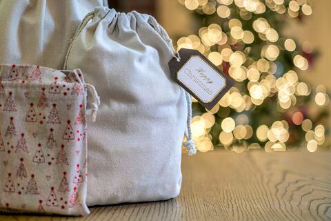 A Christmas gifts in a reusable textile bags sitting on a wooden table in front of a Christmas tree.