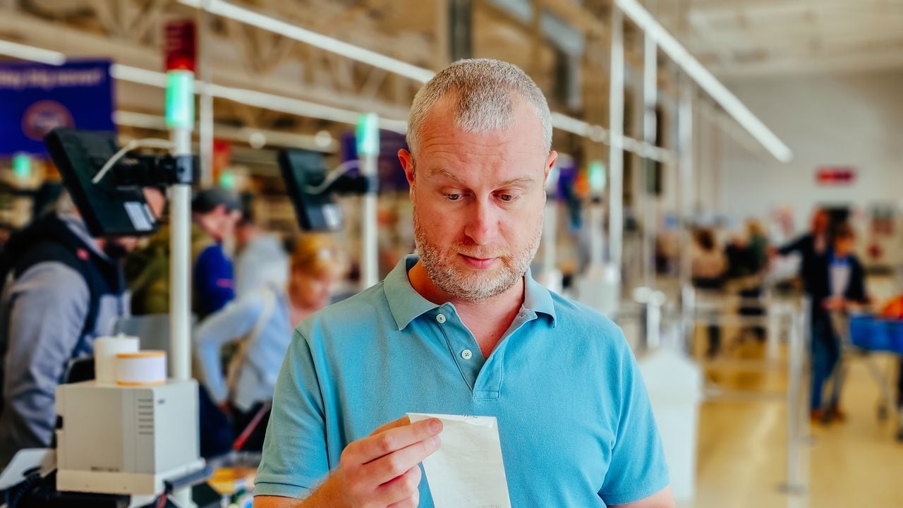 Close up color image depicting a mid adult man checking his groceries bill in the supermarket after making his purchases at the checkout. Focus on the man with the supermarket checkout defocused in the background. The man is wearing a casual blue polo shirt and has a worried expression on his face.