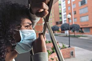 Latin man with frizzy hair and dark skin of approximately 20 years old is leaning out of the window of his house. He uses his protection mask to avoid viruses on his face. We observe a feeling of despair at the quarantine that they are going through because of the covid 19